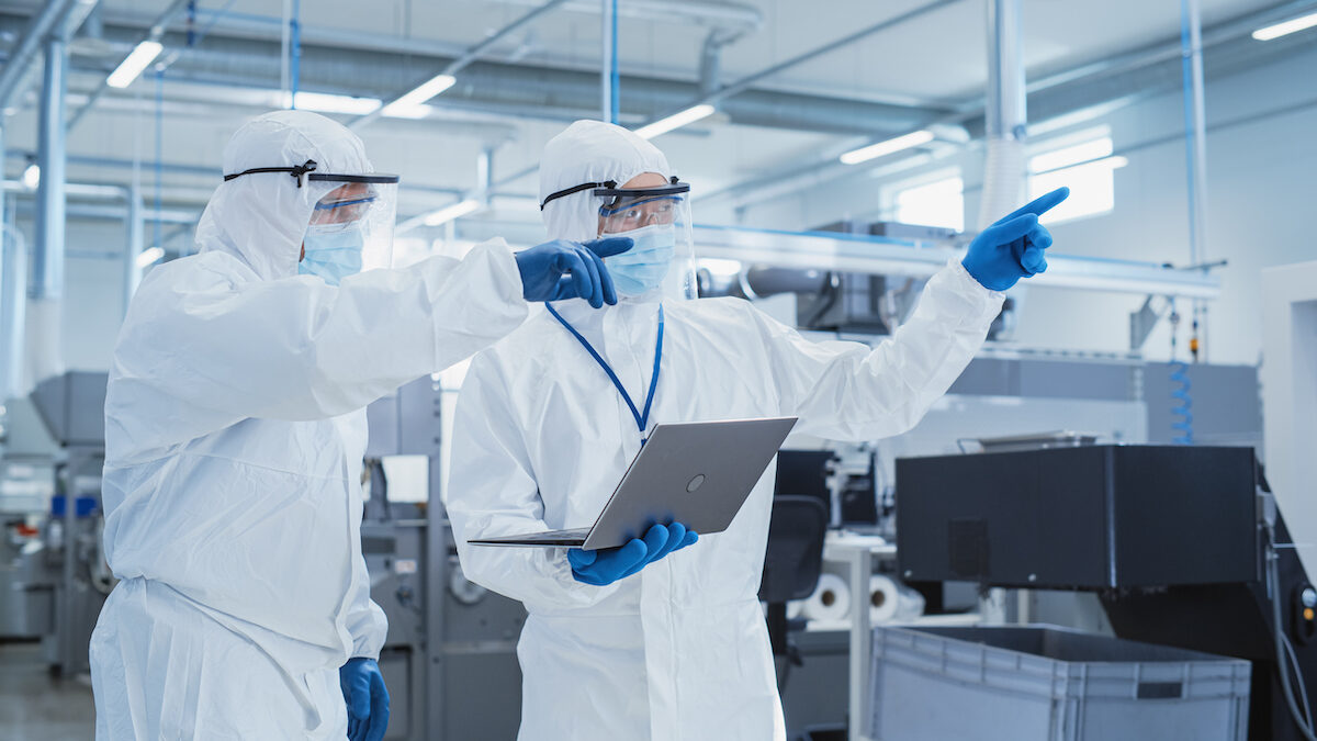 Two Scientists Standing In A Heavy Industry Factory In Sterile Coveralls And Face Masks, Using Laptop Computer. Examining Industrial Machine Settings And Configuring Production Functionality.