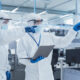 Two Scientists Standing In A Heavy Industry Factory In Sterile Coveralls And Face Masks, Using Laptop Computer. Examining Industrial Machine Settings And Configuring Production Functionality.